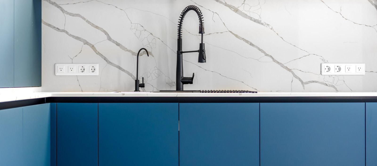 Front view of matte blue handleless cabinetry with a quartz countertop, marble vein backsplash, and tall black faucet. A clean, contemporary kitchen aesthetic.